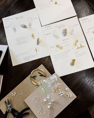 A flat lay image of jewellery pieces being assembled on a dark table. There are sketches of jewellery pieces and the unassembled parts with pliers.